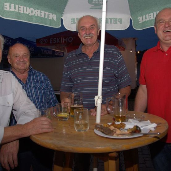 Vier Männer sitzen um einen Tisch unter einem Regenschirm. Drei von ihnen lächeln und scheinen ihr Essen und ihre Getränke zu genießen. Auf dem Tisch stehen Teller mit Essen, Gläser und Tassen. Hinter ihnen befindet sich ein Zelt mit einem Eskimo-Logo.