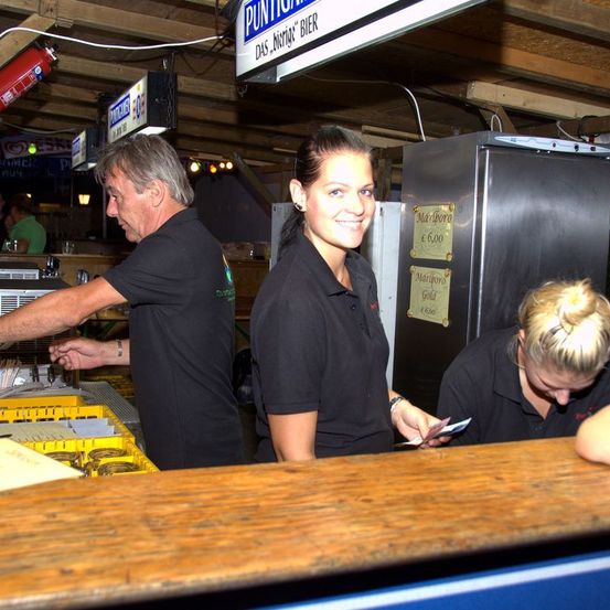 Drei Personen in schwarzen Shirts stehen an einer Bartheke. Ein Schild darüber steht 'Puntiga'. Der Mann auf der linken Seite hält einen Tablett, die Frau in der Mitte lächelt und die Frau auf der rechten Seite schaut nach unten.