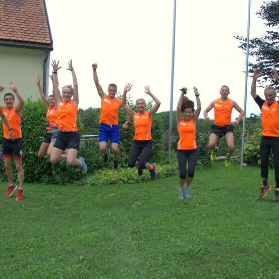 Eine Gruppe von Personen in orangefarbenen Shirts und schwarzen Shorts springt auf einem Rasenfeld mit Büschen und einem Haus dahinter in der Luft.
