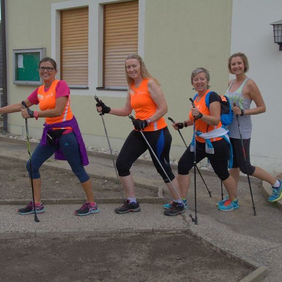 Vier Frauen in orangefarbenen Westen und mit Trekkingstöcken stehen vor einem Gebäude, lächeln und sind bereit für ein Abenteuer.
