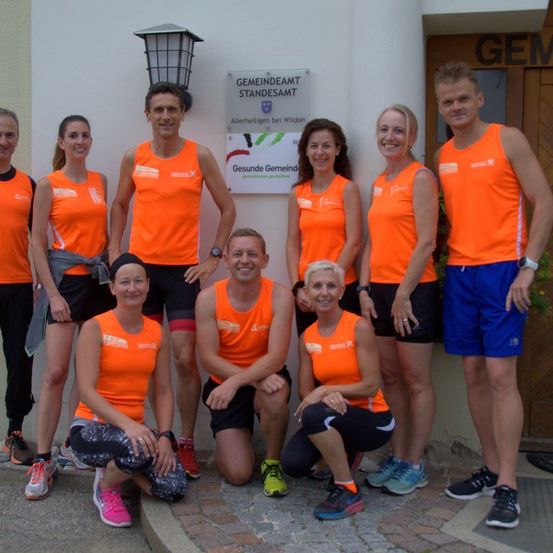 Eine Gruppe von Menschen in orangefarbenen Shirts und Turnschuhen posiert für ein Foto vor einem Gebäude. Sie lächeln und scheinen an einer Gemeinschaftsveranstaltung teilzunehmen.