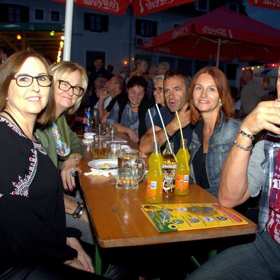 Eine Gruppe von Menschen sitzt an einem Tisch in einem Restaurant im Freien mit Getränken und Essen. Sie sind unter roten Sonnenschirmen mit Coca-Cola-Marke.