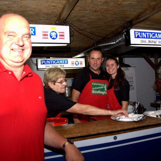 Ein Mann in einem roten Shirt und eine Frau mit Brille servieren Essen einem Mann in einem schwarzen Shirt und Schürze. Sie stehen vor einer Theke mit Tellern und Besteck.
