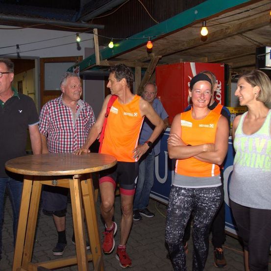 Eine Gruppe von Menschen versammelt sich nachts im Freien; ein Mann in Orange trägt eine Uhr und Turnschuhe, während andere hinter einem Tisch stehen und in der Nähe eines Coca-Cola-Kühlers stehen.