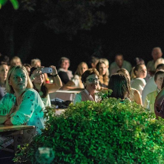 Eine Menschenmenge sitzt in einem Halbkreis und beobachtet eine Outdoor-Veranstaltung bei Nacht. Einige lächeln und machen Fotos mit ihren Handys.