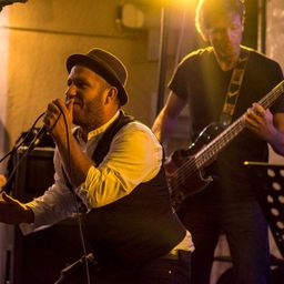 Ein Musiker mit Hut singt in ein Mikrofon, während ein anderer Musiker Gitarre spielt.