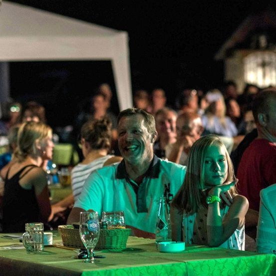 Eine dreiköpfige Familie, bestehend aus einem Mann, einem Mädchen und einer Frau, sitzt an einem Tisch mit Getränken und Essen. Sie sind von einer großen Menschenmenge umgeben, die bei Nacht unter einem Zelt sitzt.