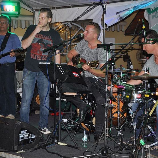 Eine Band spielt auf der Bühne, ein Mann singt in ein Mikrofon, ein anderer spielt Gitarre und ein Schlagzeuger sitzt mit Schlagzeug.