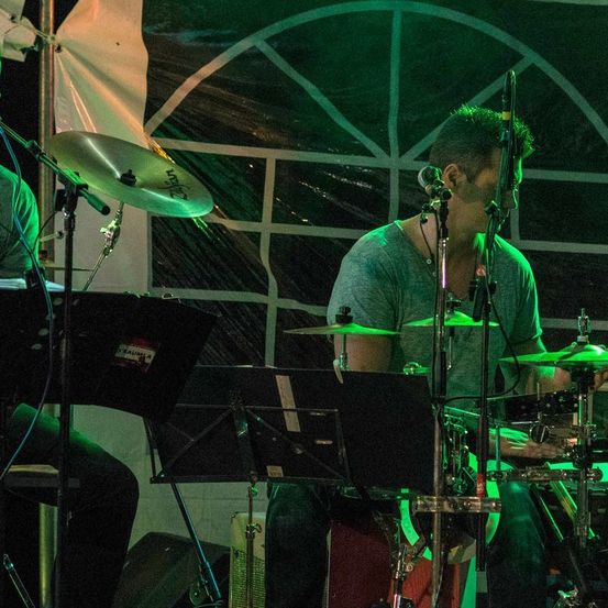 Ein Schlagzeuger spielt auf einer Bühne unter grünem Licht. Er sitzt neben einem Notenständer mit einem Becken vor ihm. Eine weitere Person sitzt zu seiner linken Seite.