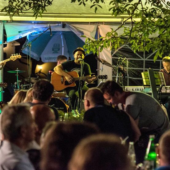 Eine Band spielt auf der Bühne, ein Gitarrist singt und spielt Gitarre, ein Pianist und ein Schlagzeuger sind ebenfalls dabei. Das Publikum sitzt davor, und ein Baum mit grünen Blättern ist über der Bühne sichtbar.