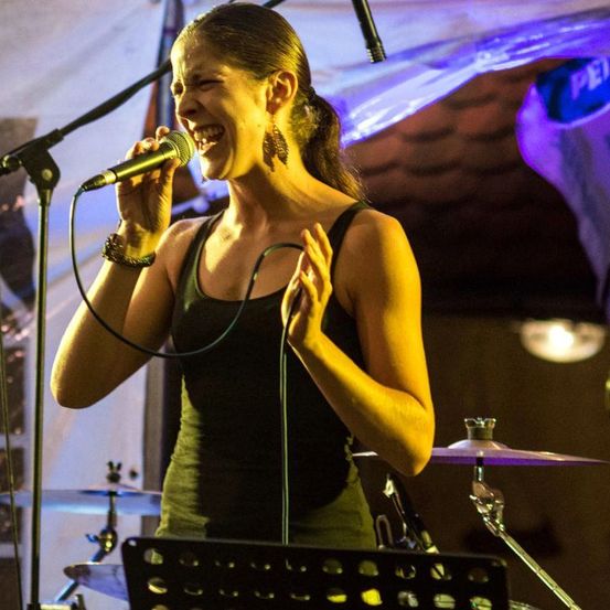 Eine Frau singt in ein Mikrofon, den Mund geöffnet, und ihre Hände erhoben. Sie trägt ein ärmelloses Oberteil und Ohrringe. Hinter ihr steht ein Schlagzeug mit Becken. Der Hintergrund ist dunkel mit lila Beleuchtung.