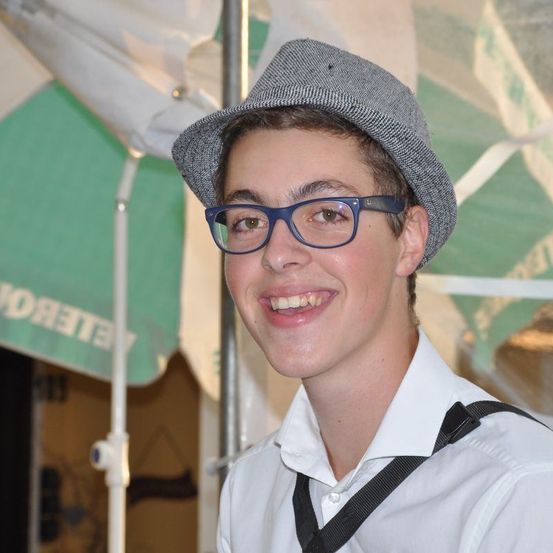 Ein junger Mann mit grauem Hut und Brille lächelt fröhlich unter einem grünen Zelt. Er hat einen schwarzen Riemen über der Schulter.