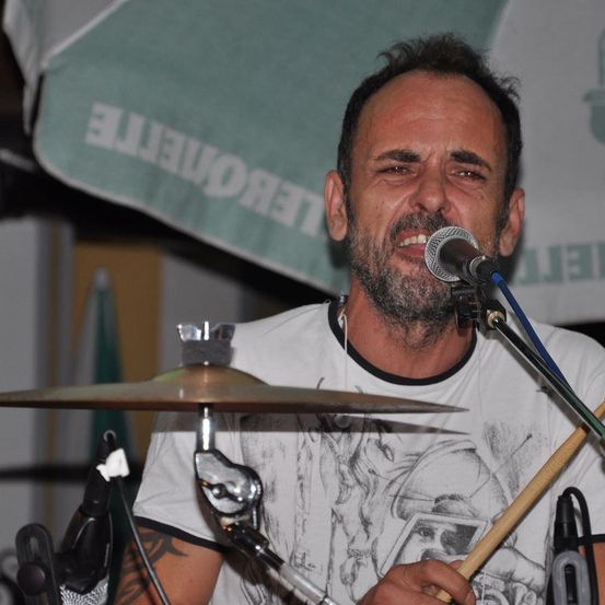 Ein Mann spielt eine Schlagzeug-Set, spielt die Becken und Drumsticks und singt in ein Mikrofon. Er trägt ein weißes T-Shirt mit einem grafischen Design.