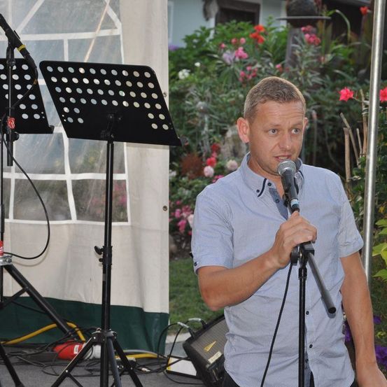 Ein Mann mit einem Mikrofon spricht in einen Ständer in einem Garten mit Blumen und einem weißen Zelt im Hintergrund.