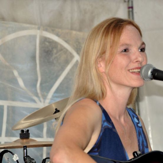 Eine Frau mit blonden Haaren tritt auf der Bühne auf, spielt Gitarre und singt in ein Mikrofon. Sie lächelt und scheint Spaß zu haben. Hinter ihr befindet sich ein Hintergrund mit einem geometrischen Design.