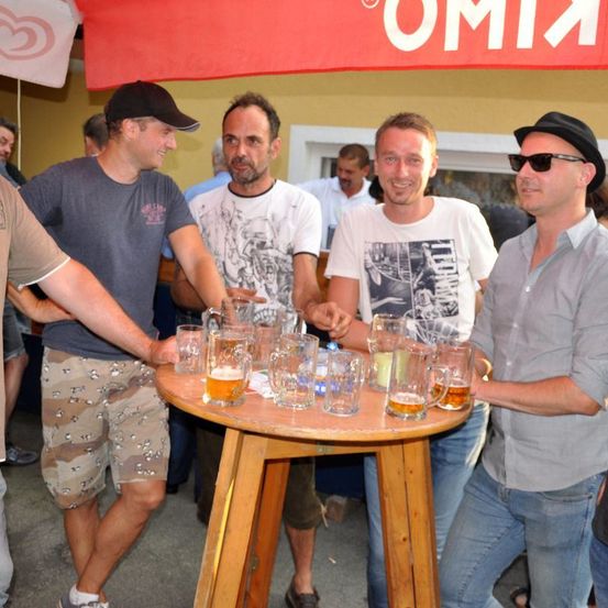 Eine Gruppe von Männern steht um einen Tisch mit mehreren Gläsern Bier. Ein Mann trägt einen schwarzen Hut und eine Sonnenbrille. Hinter ihnen ist ein Banner mit weißem Text sichtbar.