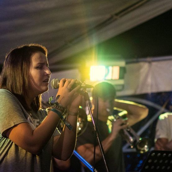 Eine Frau singt in ein Mikrofon, das von einem grünen Licht beleuchtet wird, während ein Mann hinter ihr Trompete spielt. Andere Musiker sind im Hintergrund zu sehen.