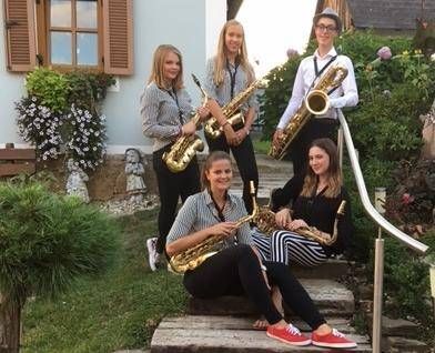 Fünf junge Frauen in gleichen Outfits posieren mit Saxophonen auf einer von Grünpflanzen umgebenen Treppe. Eine Frau sitzt auf der unteren Stufe, während die anderen hinter ihr stehen.