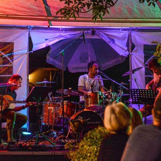 Ein nächtliches Konzert mit einer Band unter einem Zelt. Ein Mann spielt Gitarre, ein anderer singt und ein Schlagzeuger spielt dahinter. Das Publikum beobachtet es von vorne.