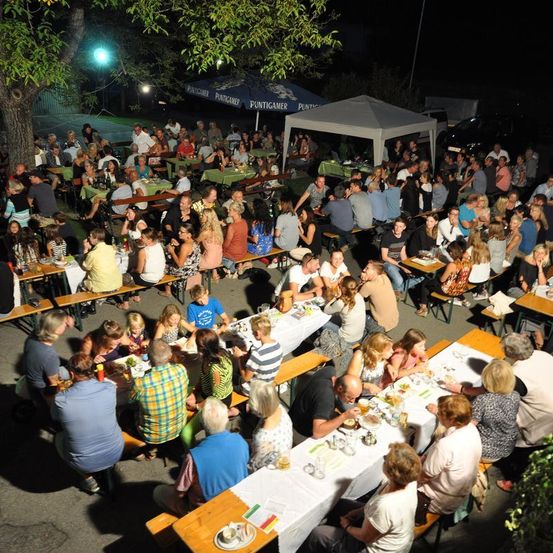 Eine große Gruppe von Menschen isst am Abend im Freien. Tische und Bänke sind aufgestellt, einige unter Zelten. Der Bereich ist beleuchtet, und die Menschen unterhalten sich und essen.