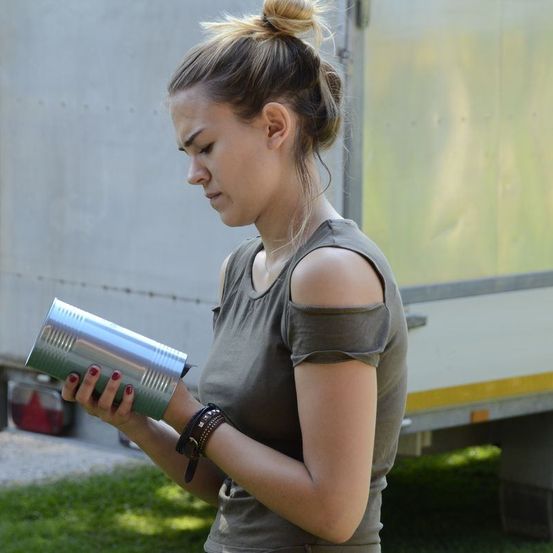 Eine Frau mit Duttfrisur und einem grauen ärmellosen Oberteil schaut sich einen Blechdosen außerhalb eines Fahrzeugs an. Sie hält die Dose mit beiden Händen.