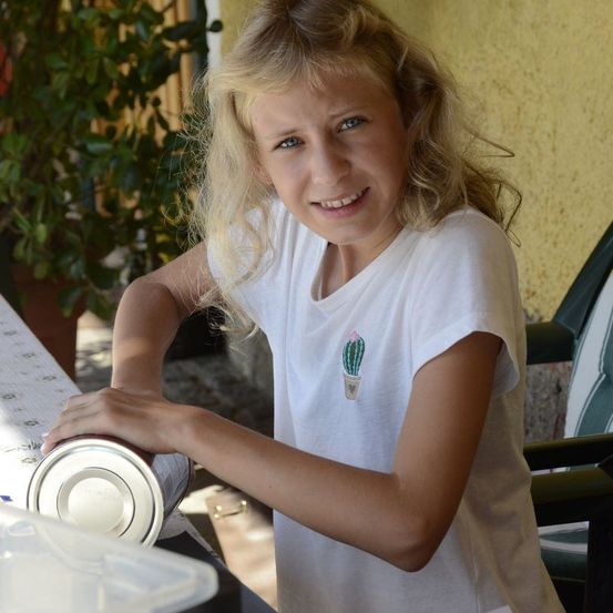 Ein junges Mädchen mit blonden Haaren hält eine Dose und lächelt, während es im Freien steht. Sie trägt ein weißes T-Shirt mit Kaktus-Aufdruck.