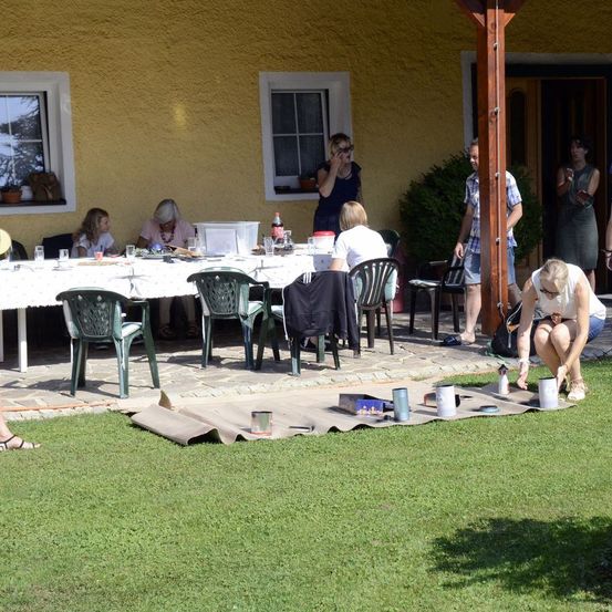 Eine Gruppe von Menschen sitzt draußen an einem Tisch, während andere in der Nähe des Eingangs stehen. Eine Person hockt auf dem Rasen.