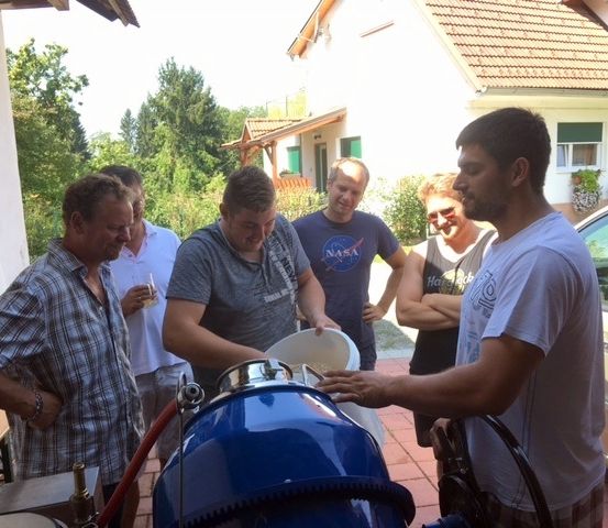 Sechs Männer versammeln sich um einen blauen Container im Freien. Ein Mann hält einen weißen Eimer. Ein anderer Mann schaut hinein. Dahinter ist ein Haus und Bäume zu sehen.