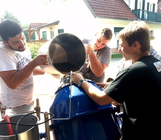 Vier Männer arbeiten zusammen in einem Garten. Einer gießt eine Substanz in einen großen blauen Fass. Dahinter befindet sich ein Haus mit Balkon.