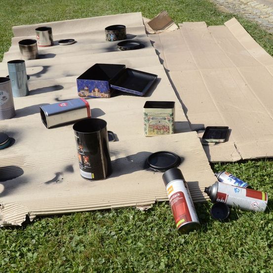 Verschiedene Metalldosen, eine Schachtel und Sprühdosen liegen auf Pappe auf dem Rasen.