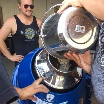 Zwei Personen füllen etwas aus einem silbernen Eimer in eine große blaue Maschine. Ein Mann in einem schwarzen Tanktop steht hinter ihnen.