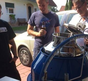 Drei Männer stehen um eine blaue tonnenförmige Maschine herum, einer hält eine Bierflasche. Der Hintergrund zeigt einen Garten mit einem Haus und einem geparkten Auto.