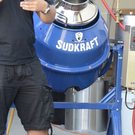 Ein Mann steht neben einem blauen Sudkraft-Mischer, trägt schwarze Kleidung und eine Armbanduhr. Der Mischer hat einen silbernen Deckel und einen roten Schlauch.