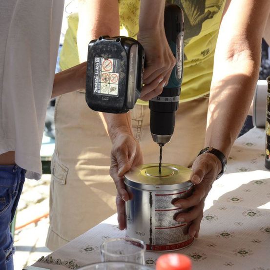 Zwei Personen benutzen eine Bohrmaschine, um in eine Dose zu bohren, während sie an einem Tisch stehen, mit einer weiteren Person in der Nähe.