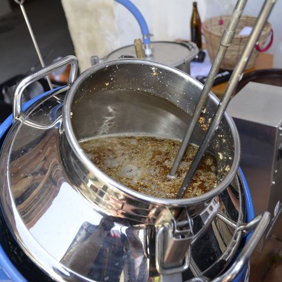 Ein Edelstahl-Braugefäß mit einem Kochlöffel, der den Inhalt rührt, reflektiert die Umgebung. Ein blauer Schlauch verbindet das Gefäß mit einem Fass, im Hintergrund sind weitere Küchenutensilien zu sehen.