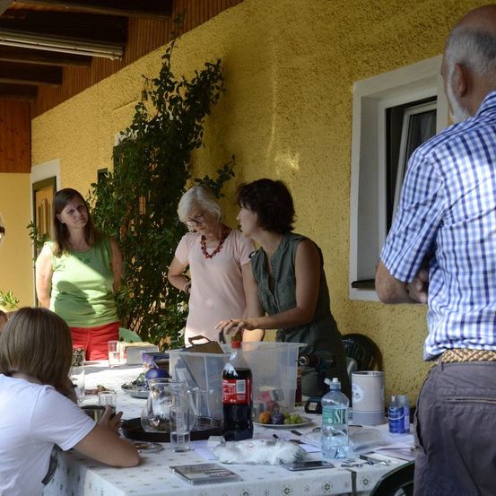 Fünf Erwachsene stehen um einen Tisch herum, auf dem verschiedene Gegenstände sind, darunter eine Flasche Coca-Cola. Eine Frau sitzt am Tisch. Die Wand ist gelb.
