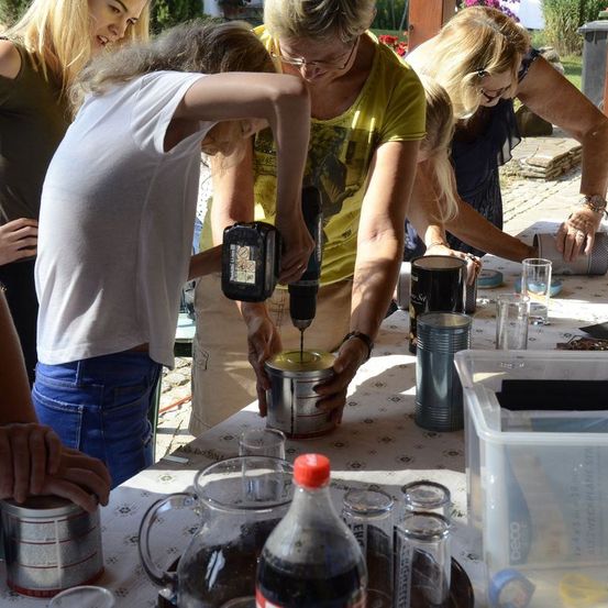 Eine Gruppe von Frauen arbeitet an einem Tisch und benutzt eine Bohrmaschine, um Dosen für ein Projekt vorzubereiten. Sie sind von verschiedenen Werkzeugen und Materialien umgeben.