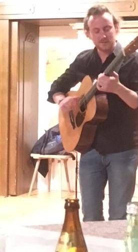 Ein Mann steht in einem Raum, hält eine Gitarre und spielt sie. Vor ihm steht eine Flasche auf einem Tisch. Hinter ihm befindet sich ein Holzstuhl und ein Beutel auf dem Boden.