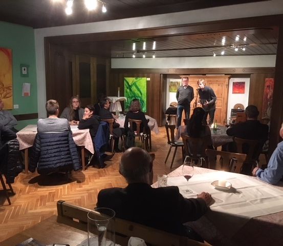 Eine Gruppe von Menschen sitzt um Tische in einem Raum. Zwei Personen stehen in der Nähe eines grünen Gemäldes. Es gibt Lampen an der Decke.