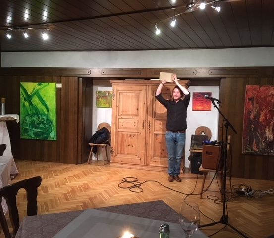 Ein Mann steht auf der Bühne und hält eine Schachtel über seinem Kopf. Er ist umgeben von Gemälden an den Wänden, einem Mikrofon und einem Tisch mit Weingläsern.