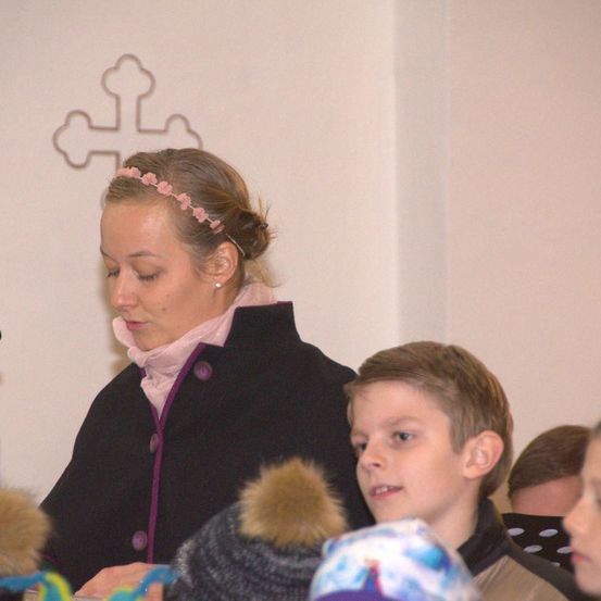 Eine Frau mit einer rosa Haarspange sitzt und sieht nach unten, möglicherweise liest sie. Ein Junge und ein Mädchen schauen in die gleiche Richtung. Sie sind wahrscheinlich in einer Kirche.