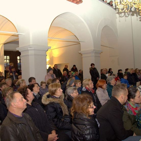Eine Gruppe von Menschen versammelt sich in einer Kirche für eine Veranstaltung. Einige tragen Brillen und Winterkleidung. Der Raum hat gewölbte Decken und weiße Wände.