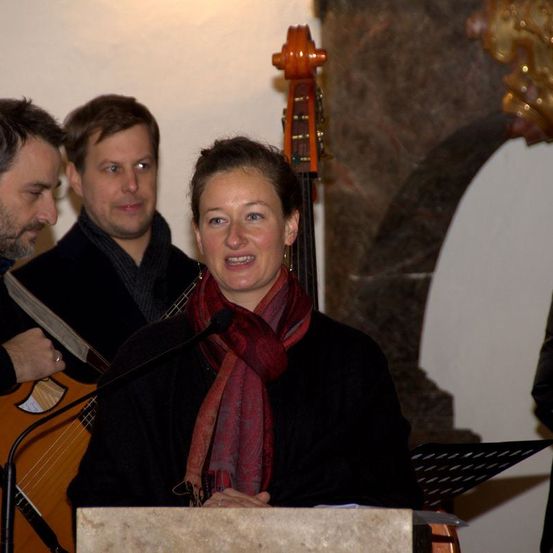 Eine Frau steht vor einem Podium und lächelt, mit einem Mikrofon vor ihr. Hinter ihr spielen zwei Männer Gitarre. Ein Mann trägt einen Schal. Die Umgebung scheint in einem Innenraum zu sein.