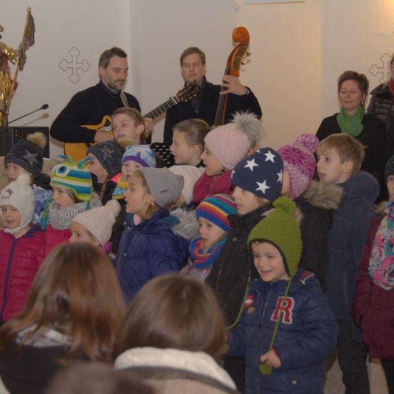 Eine Gruppe von Kindern, die Wintermützen und -jacken tragen, einige mit Schals, sind in einem Raum versammelt. Zwei Männer im Hintergrund spielen Gitarre und Cello. Eine goldene Statue befindet sich auf der linken Seite der Wand, und ein Kreuz ist auf der rechten Seite.
