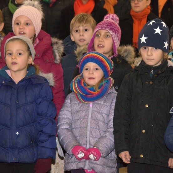 Eine Gruppe von Kindern in Winterkleidung, einige mit Mützen, stehen eng beieinander, möglicherweise bei einer öffentlichen Veranstaltung. Sie scheinen etwas zu hören.
