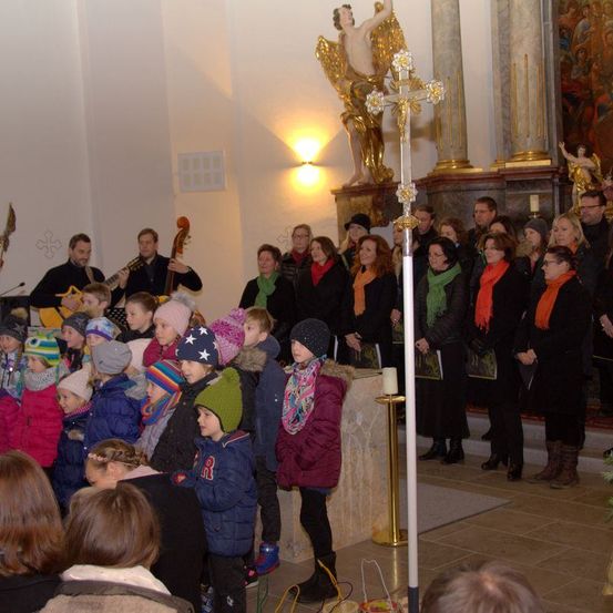 Ein Chor und eine Band spielen in einer Kirche, mit Kindern und Erwachsenen, die warm gekleidet in Mützen und Schals sind, beleuchtet von warmer Beleuchtung.