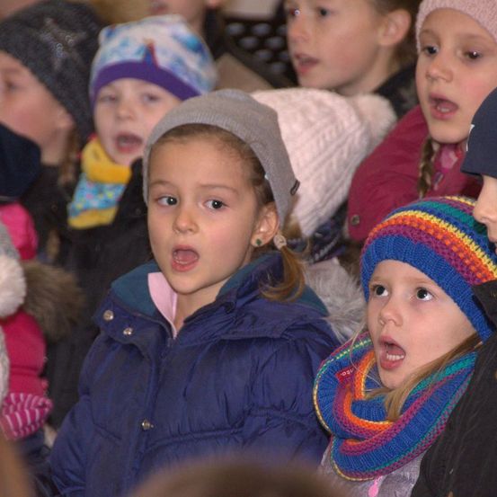 Mehrere Kinder, die Wintermützen und Schals tragen, singen laut. Sie stehen dicht beieinander. Das Mädchen vorne trägt eine blaue Jacke.