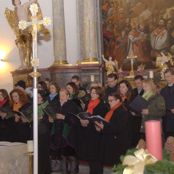 Ein Chor steht in einer Kirche mit Büchern in der Hand, einige tragen Schals und sehen ein großes Gemälde an. Eine goldene Engelsstatue steht links und eine Kerze rechts.