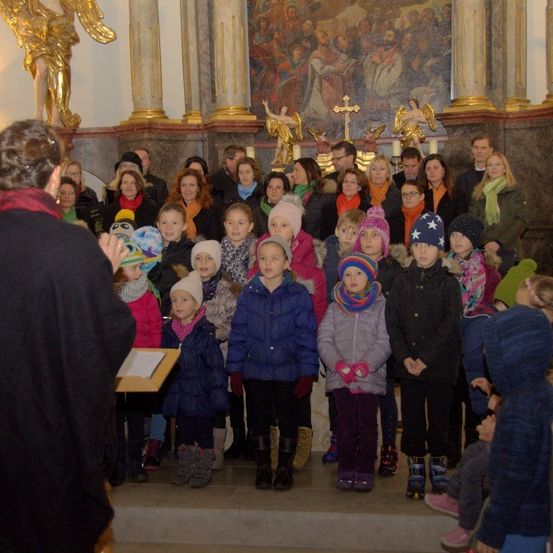 Ein Kinderchor in Winterkleidung steht vor einem Kirchenaltar, mit Erwachsenen und einer goldenen Engelstatue dahinter.
