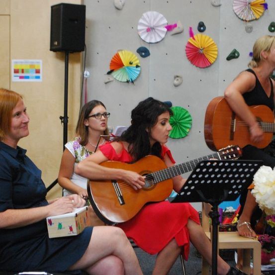 Vier Frauen spielen Gitarren in einem Raum mit einer dekorativen Wand und einem Weihnachtsbaum. Eine Frau sitzt auf einem Stuhl mit einem Notenständer.